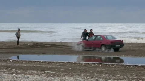 Red Iranian sedan stuck in wet sand spinning wheels on stormy polluted beach Stock Footage 321032053