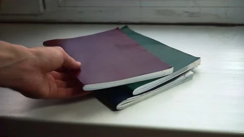Red notebook is being put on a stack of blue and green notebooks lying on the wh 스톡 동영상 168248389