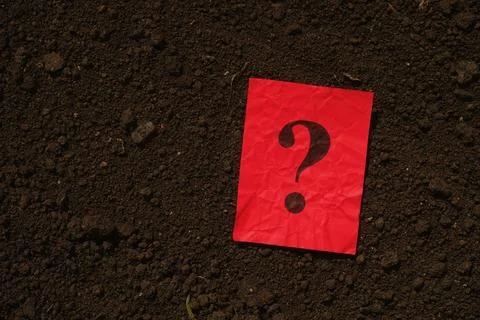 A red paper note with a question mark on it lying on a background of soil. Stock Photos