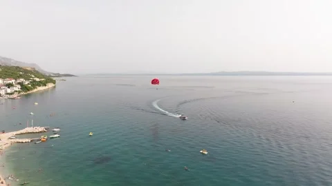 Red Parasail Gliding Past Busy Croatian Swimming Cove, Aerial Long Shot Stockbeeldmateriaal 346188997