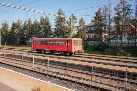 A red railcar sits on tracks bordered by tall pine trees and a residential house Stock Photos