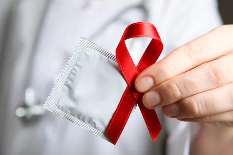 Red ribbon symbolizing AIDS and condoms in the hands of a doctor in a white coat Stock Photos
