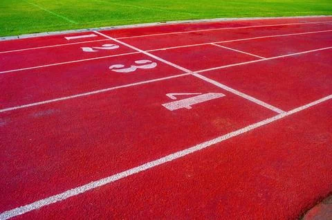 Red running track with numbered lanes beside green grass, ideal for race, s.. Stock Photos