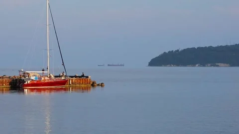Red sailboat docked at a rusty pier in a calm sea Video stock 339850697