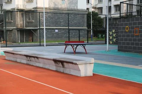 Red table tennis table beyond a long stone bench on an empty fenced recreat.. Stock Photos