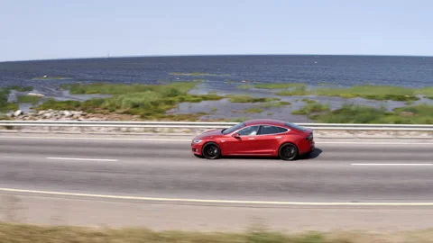 Red tesla model 3 driving down the road along the bay Stock Footage 214342762