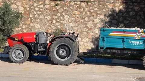 Red tractor parked beside stone wall with attached blue trailer on sunny day Stock Footage 329621825