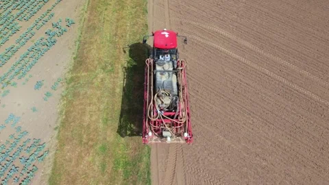 Red tractor sprays field from above. Crops line one edge, soil on other Stock Footage 345668732