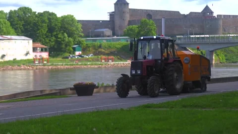 Red Tractor with Street Sweeper Driving Along River with Historic Fortress Stock Footage 337880116