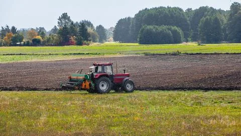 A red tractor towing a seed drill machine on a freshly plowed field with gr.. Stock Photos