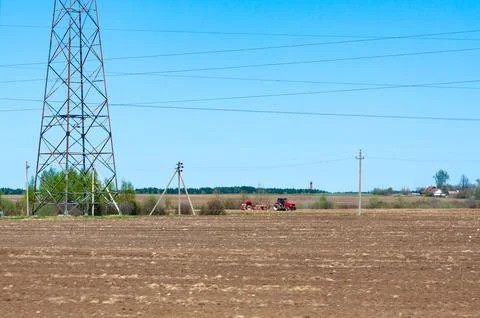 Red tractor working in a plowed soil field near power line towers under a c.. Foto stock