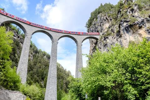 Red train crosses a stunning stone viaduct surrounded by lush green trees i.. Stock Photos