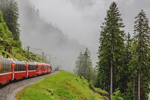 A red train winds along a narrow track amidst a misty mountain landscape Stock Photos