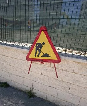 Red triangular road work hazard sign along fence in urban construction area.. Stock Photos