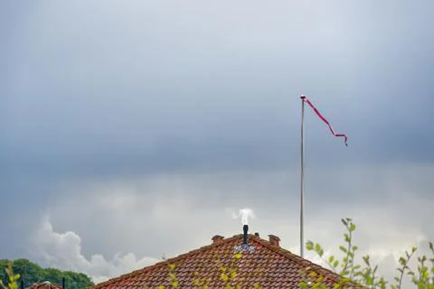 Red windsock on pole above tiled roof with smoke chimney under cloudy sky Stock Photos