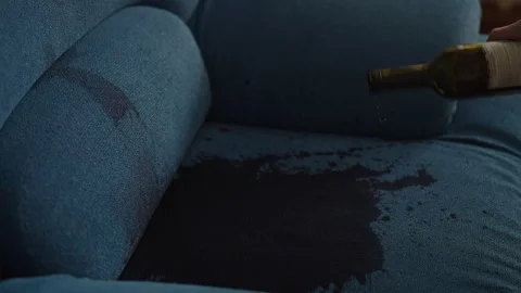 Red wine spilling from overturned bottle onto blue couch, creating large, dark Stock Footage 312375927