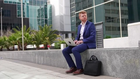 A Redhead Businessman Smiling While Working on Laptop in Urban Business Park Stock Footage 278645144