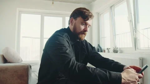 Redhead man with beard sitting at home and holding smartphone. 스톡 동영상 174716617