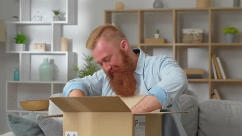 Redhead man sit on couch opens parcel box feels satisfied looking sincerely glad Stock Footage 231223735