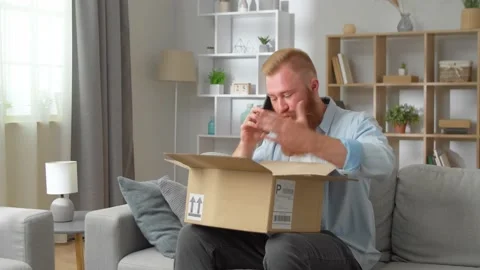 Redhead man sitting on sofa with opened parcel express complaints to seller or Vidéo 231223698