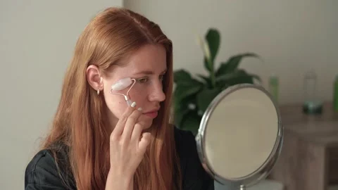 Redhead woman using jade roller on face looking in the mirror Stock Footage 199262129