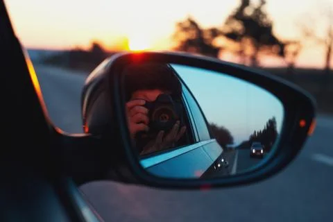 Reflection in car mirror of young guy taking photo of sunset with camera. Stock Photos