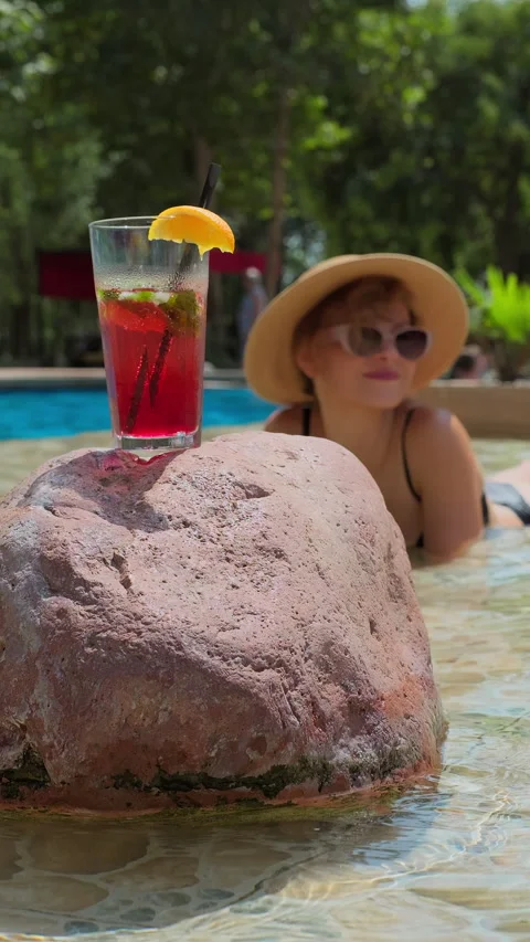 Refreshing summer cocktail with an orange slice rests on a rock near a pool Stock Footage 313742545