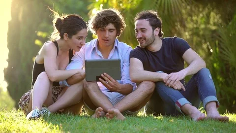 Relaxed and  happy friends using tablet sitting on meadow in summer time Stock Footage 76401458