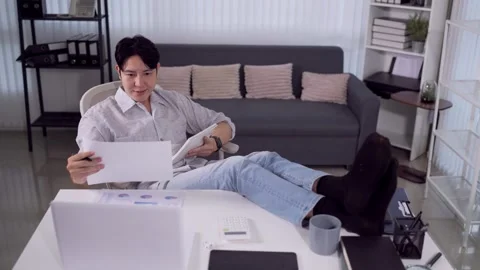 Relaxed Asian man reviewing documents with feet on desk in casual home offi.. Stock Footage 345156702