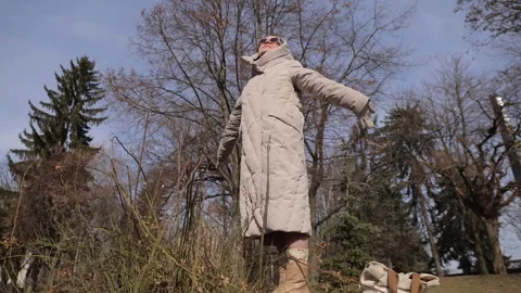 Relaxed elderly woman in jacket with spread arms looks at sun in spring park Video stock 329465701