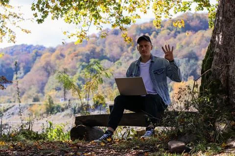 Relaxed guy gesturing wave at camera with laptop during outdoor autumn work 스톡 사진
