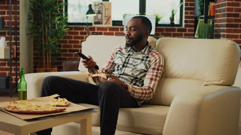 Relaxed guy taking bite of slice of pizza in living room Stock Footage 232991520