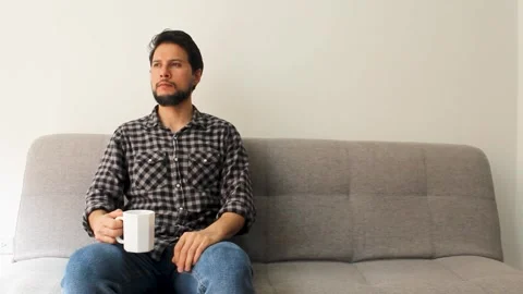 A relaxed hispanic male sitting at the left side of a couch drinking a beverage Vídeo Stock 340583397
