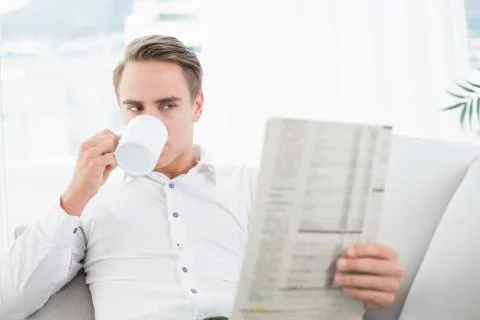 Relaxed man drinking coffee while reading newspaper on sofa Stock Photos