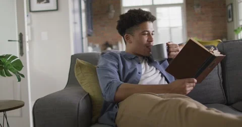 Relaxed man sitting on sofa in living room reading book and drinking coffee 動画素材 339160147