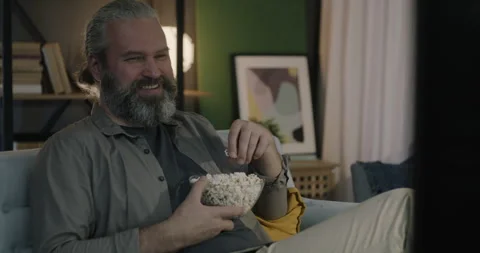 Relaxed middle-aged man watching funny comedy on TV laughing eating popcorn 動画素材 253477382