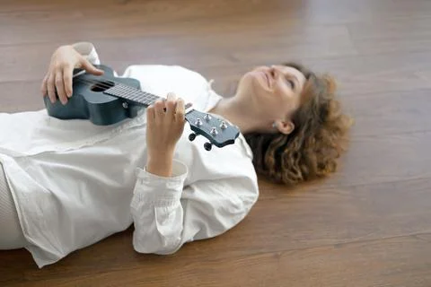 A relaxed person playing a ukulele while lying on a wooden floor in a cozy Stock Photos