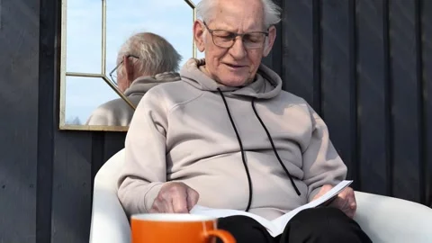 Relaxed senior reading book while sitting in white chair, drinking from orange Stock Footage 306924846