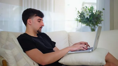 Relaxed serious millennial guy student freelancer using laptop on couch at home. Stock Footage 276978418