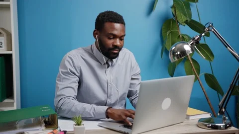 Relaxed smiling young Afro man with headphones using laptop in a bright office Stock Footage 108170992