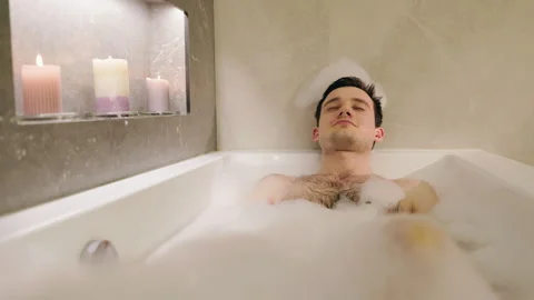 Relaxed young man enjoying a bubble bath with lit candles in a cozy bathroom. Stock Footage 309645124