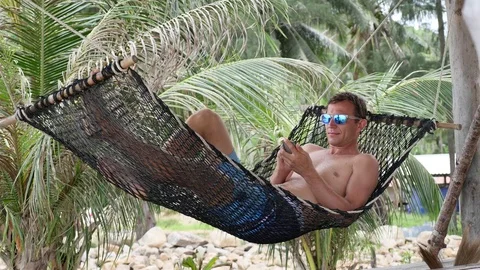 Relaxed Young Man Looking at Mobile Phone in Hammock at the Beach near Sea Stock Footage 74684467