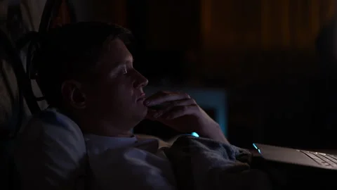 Relaxed young man lying in bed at night in dark bedroom, illuminated by screen Stock Footage 284873106