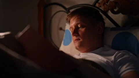 Relaxed young man lying in bed reading book in warm light, creating cozy, serene Video stock 284873194