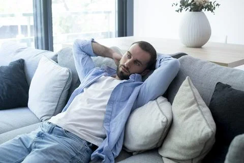 A relaxed young man lying on a gray sofa in a bright living room, casually .. Stock Photos