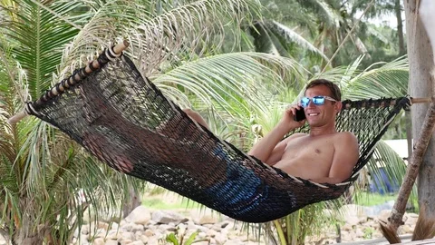 Relaxed Young Man Talking on Mobile Phone in Hammock at the Beach near Sea Stock Footage 74684504