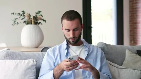 Relaxed young man using smartphone while sitting on couch at home Stock Footage 335588162