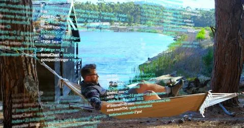 Relaxing bespectacled coder in hammock at lakeside wooden cabin coding on laptop Foto stock