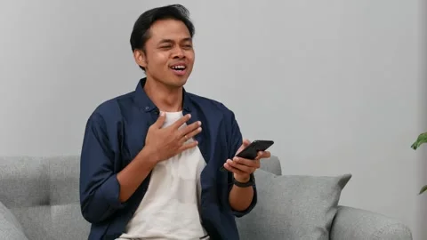 Relieved Young Asian man celebrating victory while using mobile phone at home Stock Footage 332762270