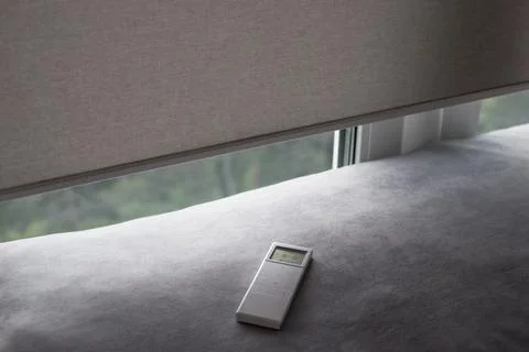 Remote control lies on a gray surface next to partially opened window blinds. Stock Photos